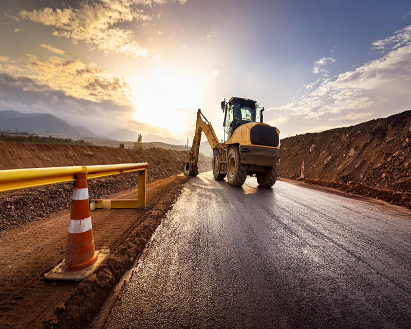 A construction site with a road grader working on a newly paved road, surrounded by dirt mounds and safety cones, under a cloudy sky during sunset.