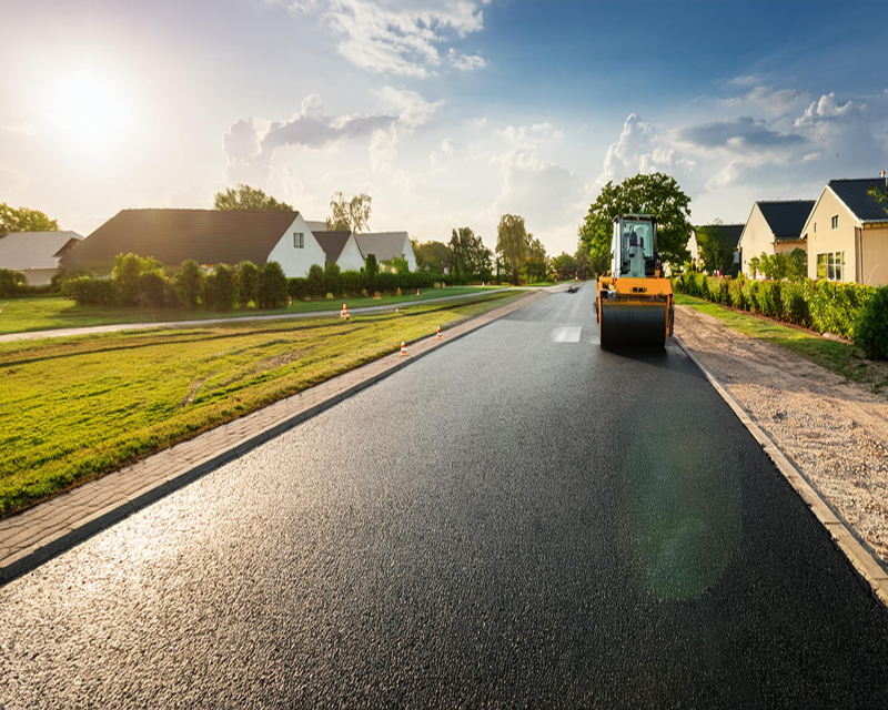 A newly marked road with double white lines in a suburban area, with houses and greenery on either side, under a clear blue sky.